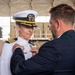 A military promotion or award ceremony is taking place, in which a civilian man is pinning a badge or insignia onto the uniform of a U.S. Navy officer. The officer is wearing a white dress uniform with gold shoulder boards indicating a senior rank (Captain or Commander), a white combination cover with gold scrambled eggs, and service ribbons. The setting appears to be a military hangar or pier facility.