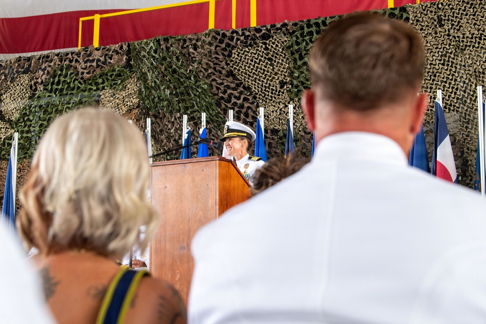 A military officer in white dress uniform speaks at a podium during what appears to be a formal ceremony. The setting is decorated with camouflage netting and multiple flags, suggesting a military change of command or similar official event. Two audience members are visible in the foreground, viewed from behind.