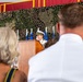 A military officer in white dress uniform speaks at a podium during what appears to be a formal ceremony. The setting is decorated with camouflage netting and multiple flags, suggesting a military change of command or similar official event. Two audience members are visible in the foreground, viewed from behind.