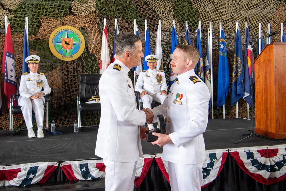 A formal U.S. Navy military ceremony is taking place, likely a change of command or award presentation. Two naval officers in white dress uniforms are shaking hands while exchanging what appears to be a plaque or award in the foreground. The event is staged under camouflage netting with multiple U.S. state and military flags displayed on a decorated platform.