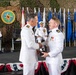 A formal U.S. Navy military ceremony is taking place, likely a change of command or award presentation. Two naval officers in white dress uniforms are shaking hands while exchanging what appears to be a plaque or award in the foreground. The event is staged under camouflage netting with multiple U.S. state and military flags displayed on a decorated platform.