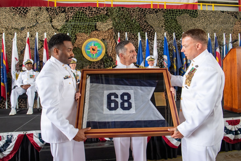 A military ceremony involving U.S. Navy personnel in dress white uniforms, where a framed pennant flag bearing the number '68' is being presented. A seal visible in the background reads 'Commander Task Force' with the designation 'CTF 68'. The event appears to be a change of command or deactivation ceremony held in an indoor venue decorated with camouflage netting, flags, and patriotic bunting.