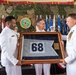 A military ceremony involving U.S. Navy personnel in dress white uniforms, where a framed pennant flag bearing the number '68' is being presented. A seal visible in the background reads 'Commander Task Force' with the designation 'CTF 68'. The event appears to be a change of command or deactivation ceremony held in an indoor venue decorated with camouflage netting, flags, and patriotic bunting.