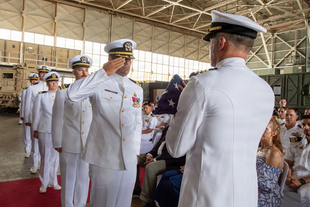 A formal U.S. Navy military ceremony is taking place inside a large hangar facility. Officers in white dress uniforms are participating in what appears to be a change of command or retirement ceremony, with one officer saluting while another holds a folded American flag. Seated guests and additional uniformed personnel observe the proceedings.