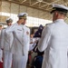 A formal U.S. Navy military ceremony is taking place inside a large hangar facility. Officers in white dress uniforms are participating in what appears to be a change of command or retirement ceremony, with one officer saluting while another holds a folded American flag. Seated guests and additional uniformed personnel observe the proceedings.