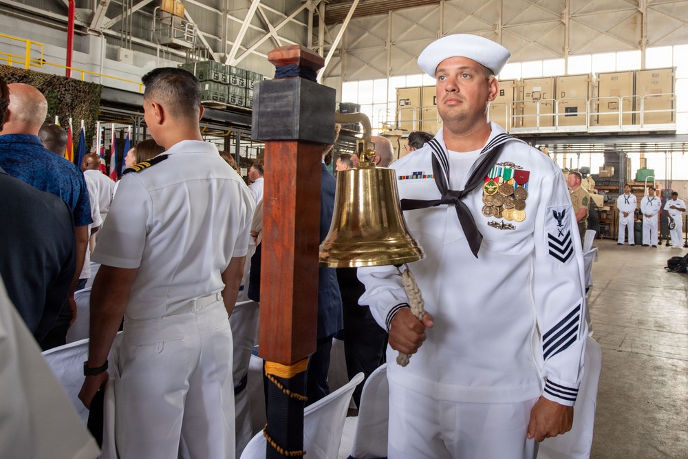 A U.S. Navy sailor in dress white uniform holds a brass ship's bell during what appears to be a formal military ceremony held inside a large hangar or warehouse facility. The sailor displays multiple service ribbons, medals, and rating badges indicating significant service experience. Several other naval personnel and civilians are visible in the background attending the ceremony.