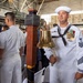 A U.S. Navy sailor in dress white uniform holds a brass ship's bell during what appears to be a formal military ceremony held inside a large hangar or warehouse facility. The sailor displays multiple service ribbons, medals, and rating badges indicating significant service experience. Several other naval personnel and civilians are visible in the background attending the ceremony.