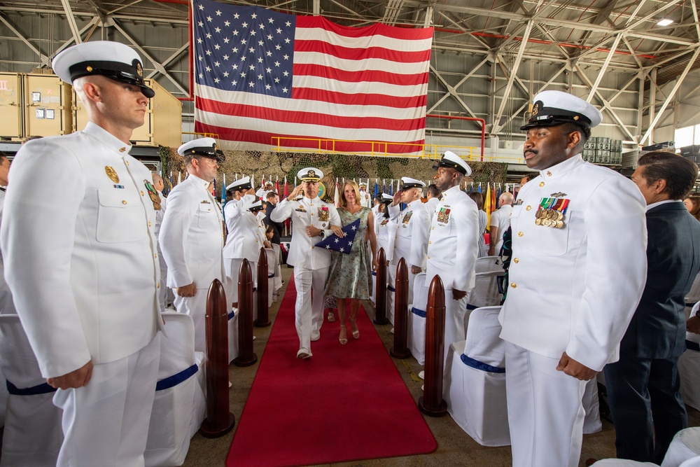 A formal U.S. Navy military ceremony is taking place inside a large hangar or warehouse facility. A naval officer in dress whites escorts a civilian woman down a red carpet aisle, carrying a folded American flag, while personnel render salutes on both sides. A large American flag is prominently displayed in the background, suggesting this is a change of command, retirement, or commissioning ceremony.