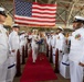 A formal U.S. Navy military ceremony is taking place inside a large hangar or warehouse facility. A naval officer in dress whites escorts a civilian woman down a red carpet aisle, carrying a folded American flag, while personnel render salutes on both sides. A large American flag is prominently displayed in the background, suggesting this is a change of command, retirement, or commissioning ceremony.