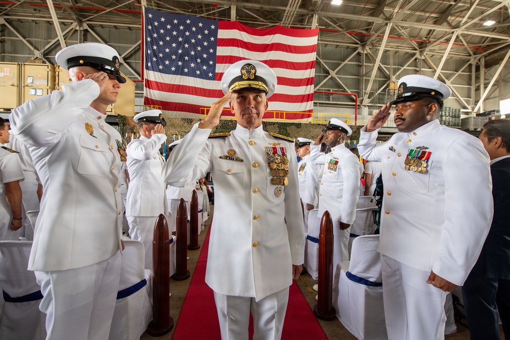 A formal U.S. Navy military ceremony is taking place inside a large hangar facility. A senior officer, prominently centered on a red carpet and adorned with numerous medals and gold insignia, is rendering a salute while flanked by rows of sailors in white dress uniforms also saluting. A large American flag is prominently displayed in the background.