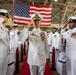 A formal U.S. Navy military ceremony is taking place inside a large hangar facility. A senior officer, prominently centered on a red carpet and adorned with numerous medals and gold insignia, is rendering a salute while flanked by rows of sailors in white dress uniforms also saluting. A large American flag is prominently displayed in the background.
