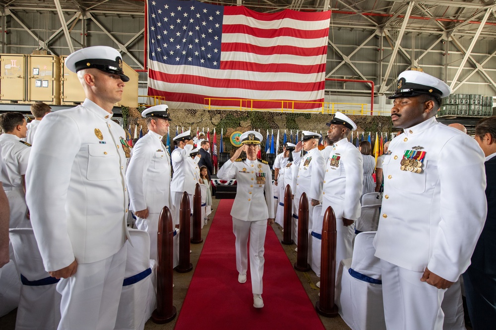 A formal U.S. Navy change of command or commissioning ceremony taking place inside a large military hangar. A senior officer in white dress uniform walks down a red carpet aisle while flanked by saluting personnel in white dress uniforms. A large American flag is prominently displayed in the background alongside multiple military unit flags.