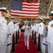 A formal U.S. Navy change of command or commissioning ceremony taking place inside a large military hangar. A senior officer in white dress uniform walks down a red carpet aisle while flanked by saluting personnel in white dress uniforms. A large American flag is prominently displayed in the background alongside multiple military unit flags.