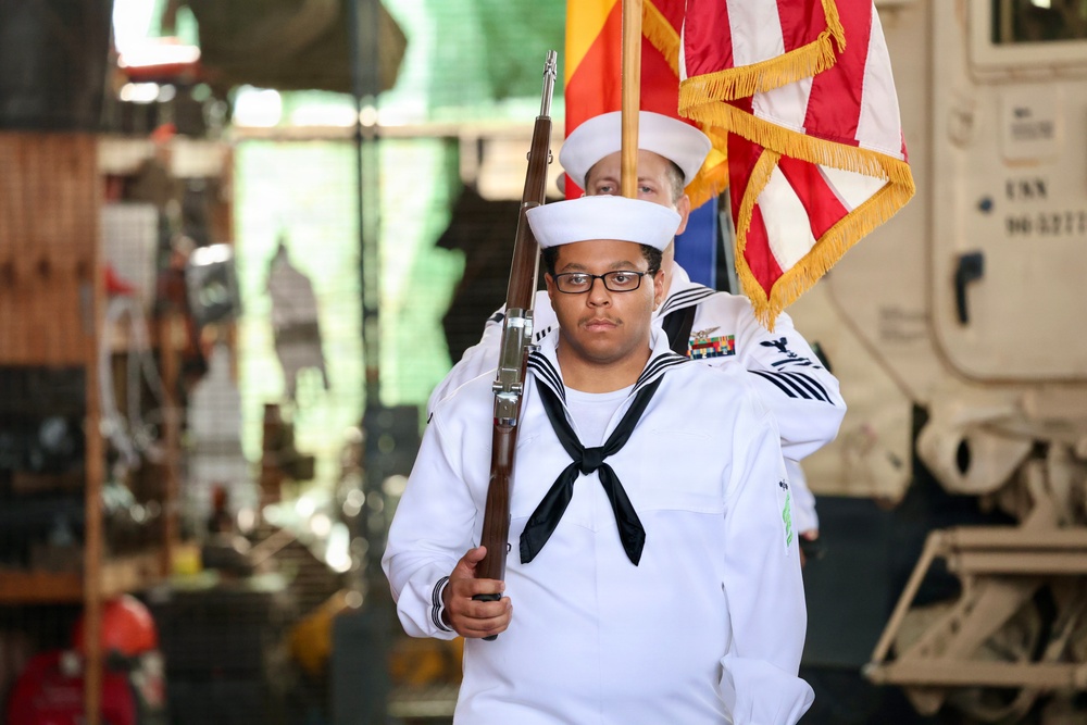 A U.S. Navy sailor in dress white uniform carries a rifle while participating in a color guard ceremony. The sailor is in the foreground, wearing ribbons and a Petty Officer insignia, with a second sailor visible behind carrying the American flag with gold fringe. The setting appears to be an indoor or covered venue with military vehicles visible in the background.