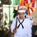 A U.S. Navy sailor in dress white uniform carries a rifle while participating in a color guard ceremony. The sailor is in the foreground, wearing ribbons and a Petty Officer insignia, with a second sailor visible behind carrying the American flag with gold fringe. The setting appears to be an indoor or covered venue with military vehicles visible in the background.