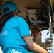 Two individuals are inside what appears to be a military vehicle, likely a HUMVEE or similar tactical vehicle. One person wearing a teal/turquoise t-shirt and matching cap is seated with their back to the camera, while a second person wearing an olive/military-style cap looks up and appears to be explaining or demonstrating something. The interior shows complex wiring, control panels, and mechanical components typical of military vehicles.