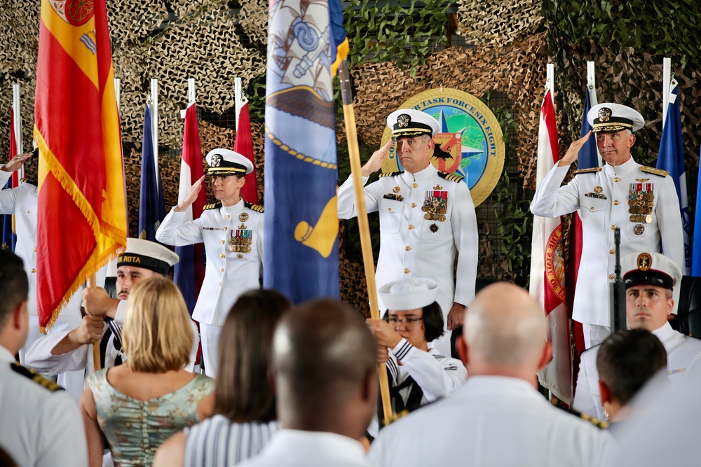 A formal military change of command or awards ceremony is taking place, with three senior U.S. Navy officers in white dress uniforms rendering salutes on a stage. The backdrop features camouflage netting, multiple national and military flags, and a seal reading 'Commander Task Force Six Eight.' Attendees including color guard members and civilians are visible in the foreground.