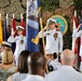 A formal military change of command or awards ceremony is taking place, with three senior U.S. Navy officers in white dress uniforms rendering salutes on a stage. The backdrop features camouflage netting, multiple national and military flags, and a seal reading 'Commander Task Force Six Eight.' Attendees including color guard members and civilians are visible in the foreground.