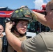 A firefighter instructor is fitting a structural firefighting helmet onto a trainee in front of a red fire apparatus. The trainee is wearing tan firefighting gear with a U.S. flag patch visible on the sleeve. A third person in similar tan attire is visible in the background observing the scene.