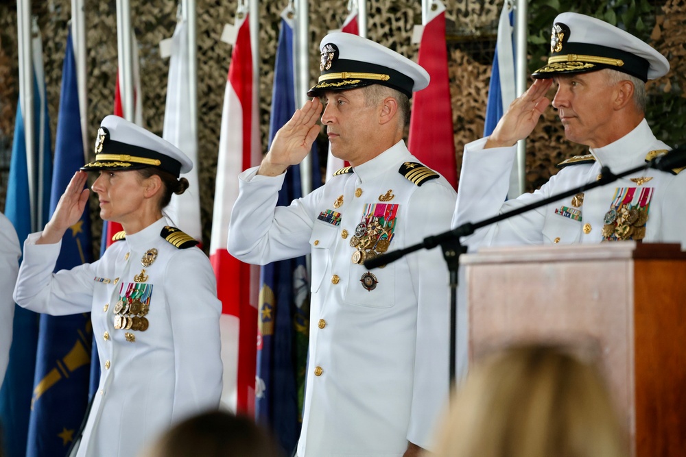 Three U.S. Navy officers in white dress uniforms are simultaneously rendering a salute during what appears to be a formal military ceremony. The setting includes multiple flags in the background and a wooden podium with a microphone, suggesting a change of command or similar official event. The officers display extensive medal and ribbon collections indicating senior rank and long service.