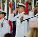 Three U.S. Navy officers in white dress uniforms are simultaneously rendering a salute during what appears to be a formal military ceremony. The setting includes multiple flags in the background and a wooden podium with a microphone, suggesting a change of command or similar official event. The officers display extensive medal and ribbon collections indicating senior rank and long service.