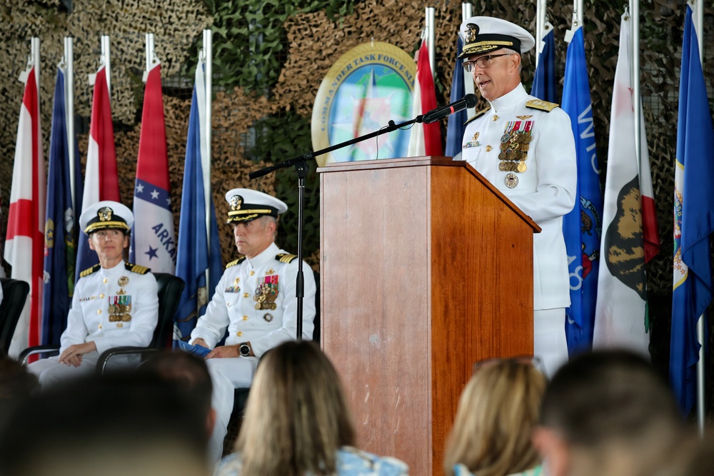 A formal U.S. Navy military ceremony is taking place, likely a change of command or similar official event. A senior naval officer in white dress uniform is speaking at a podium, while two other uniformed officers are seated behind him. Multiple U.S. state flags and other flags are displayed in the background, along with a 'Commander Task Force' emblem.