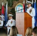 A formal U.S. Navy military ceremony is taking place, likely a change of command or similar official event. A senior naval officer in white dress uniform is speaking at a podium, while two other uniformed officers are seated behind him. Multiple U.S. state flags and other flags are displayed in the background, along with a 'Commander Task Force' emblem.