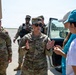 U.S. Army soldiers in OCP (Operational Camouflage Pattern) uniforms are interacting with civilian cadets on what appears to be a military airfield or tarmac. A central female soldier, identifiable by her U.S. Army tape and staff sergeant rank, appears to be briefing or instructing two young women wearing teal 'CADET' baseball caps. A CH-47 Chinook helicopter is visible in the background.