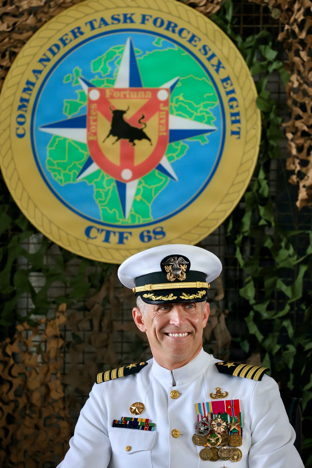 A senior U.S. Navy officer in white dress uniform poses formally in front of the Commander Task Force Six Eight (CTF 68) insignia. The officer displays multiple medals, ribbons, and insignia consistent with a high-ranking naval captain or flag officer. The background features camouflage netting with the official CTF 68 emblem prominently displayed.