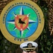 A senior U.S. Navy officer in white dress uniform poses formally in front of the Commander Task Force Six Eight (CTF 68) insignia. The officer displays multiple medals, ribbons, and insignia consistent with a high-ranking naval captain or flag officer. The background features camouflage netting with the official CTF 68 emblem prominently displayed.