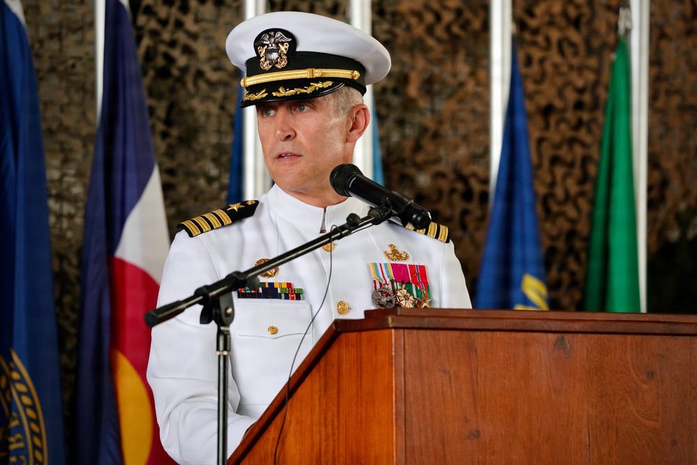 A U.S. Navy officer in white dress uniform is speaking at a podium with a microphone. The setting appears to be a formal military ceremony, indicated by the presence of multiple flags in the background and camouflage netting. The officer displays multiple service ribbons and medals on his uniform.