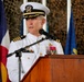 A U.S. Navy officer in white dress uniform is speaking at a podium with a microphone. The setting appears to be a formal military ceremony, indicated by the presence of multiple flags in the background and camouflage netting. The officer displays multiple service ribbons and medals on his uniform.
