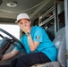 A young woman wearing a white baseball cap and a blue t-shirt with a Romanian flag patch is seated in the driver's seat of a fire truck. She is laughing and covering her mouth with her hand in an amused or surprised expression. The vehicle's red exterior, steering wheel, and gray interior are clearly visible.