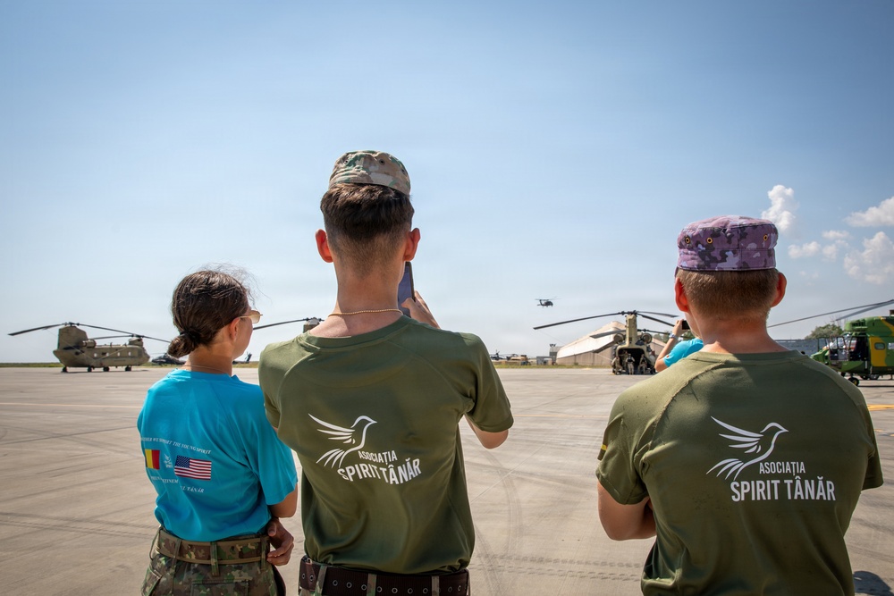 Three individuals wearing 'Asociația Spirit Tânăr' (Young Spirit Association) shirts stand on a military airfield tarmac observing CH-47 Chinook helicopters. Two of the individuals are photographing the aircraft with mobile phones. The scene appears to be a military open day or youth program visit at a Romanian air base, indicated by the Romanian and US flags visible on one shirt.