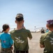 Three individuals wearing 'Asociația Spirit Tânăr' (Young Spirit Association) shirts stand on a military airfield tarmac observing CH-47 Chinook helicopters. Two of the individuals are photographing the aircraft with mobile phones. The scene appears to be a military open day or youth program visit at a Romanian air base, indicated by the Romanian and US flags visible on one shirt.