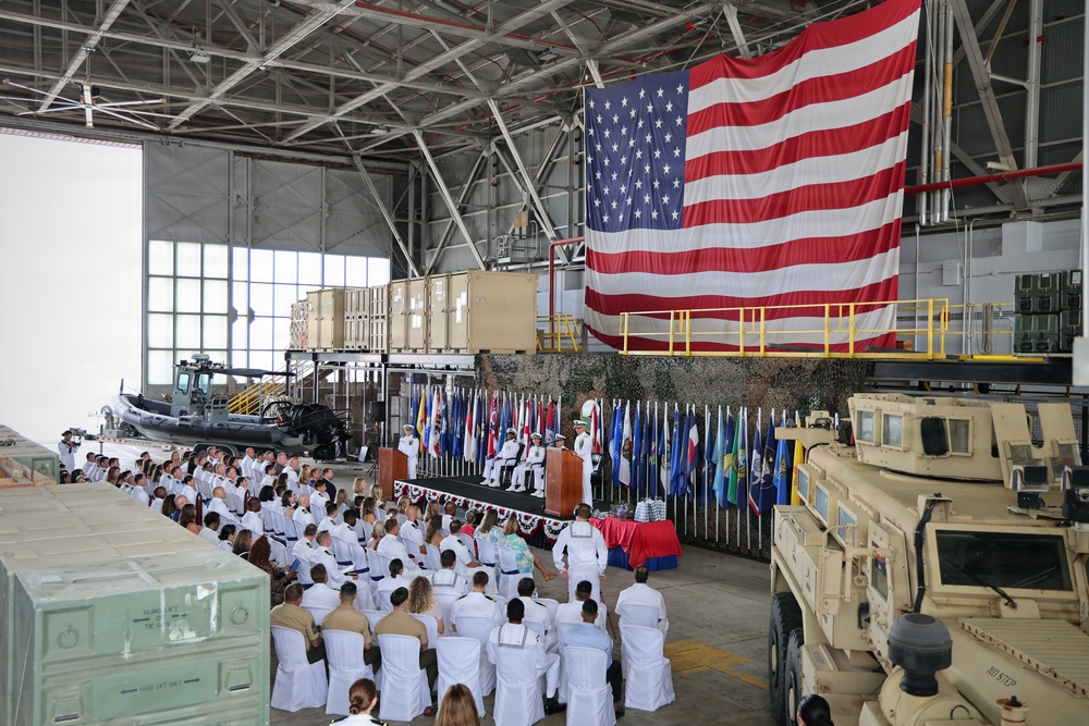 A formal military ceremony is taking place inside a large hangar facility, with personnel in white dress uniforms seated in rows facing a stage. A large American flag is prominently displayed as a backdrop behind the podium, flanked by numerous national and unit flags. Military equipment including a rigid inflatable boat, armored vehicles (MRAP), and cargo crates are visible throughout the hangar.