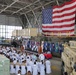 A formal military ceremony is taking place inside a large hangar facility, with personnel in white dress uniforms seated in rows facing a stage. A large American flag is prominently displayed as a backdrop behind the podium, flanked by numerous national and unit flags. Military equipment including a rigid inflatable boat, armored vehicles (MRAP), and cargo crates are visible throughout the hangar.
