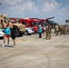 An outdoor public event, likely a military or emergency services open day, taking place on a tarmac or airfield. Various vehicles including a military MRAP/JLTV, police car, and red fire trucks are on display. Civilians wearing matching teal/turquoise shirts interact with uniformed military personnel.