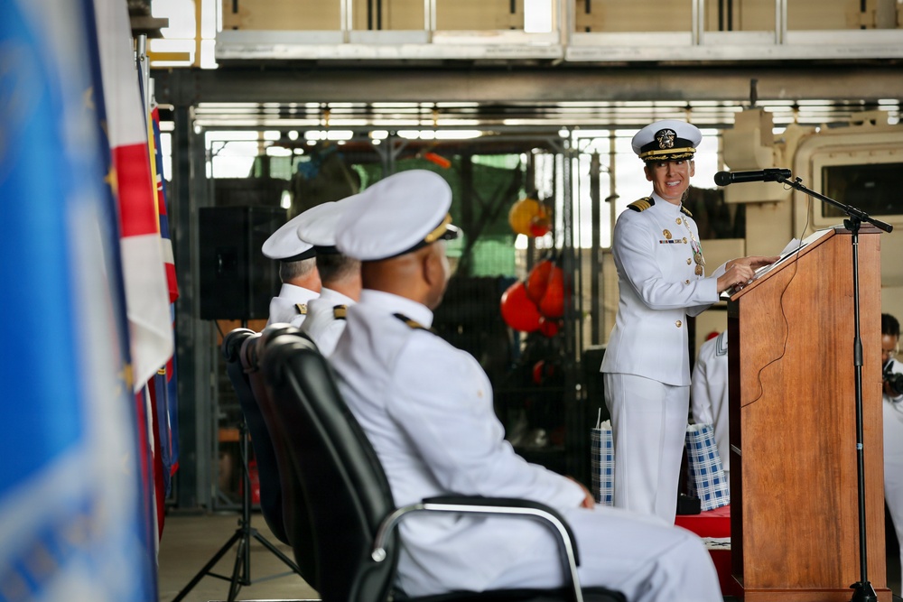 A female naval officer in white dress uniform is delivering a speech at a podium with a microphone during what appears to be a formal military ceremony. Two other naval officers in white uniforms are seated in the foreground facing the speaker. The event appears to be taking place in a ship's hangar bay or similar naval facility.