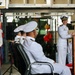 A female naval officer in white dress uniform is delivering a speech at a podium with a microphone during what appears to be a formal military ceremony. Two other naval officers in white uniforms are seated in the foreground facing the speaker. The event appears to be taking place in a ship's hangar bay or similar naval facility.