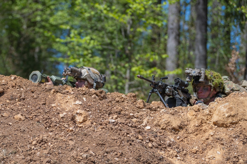 Two soldiers in full combat gear are positioned behind a dirt berm in a wooded area, taking a defensive or observational posture. One soldier is aiming a light machine gun equipped with a bipod and optic, while the other appears to be using a handheld device. The setting and equipment suggest a military training exercise or tactical field operation.