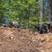 Two soldiers in full combat gear are positioned behind a dirt berm in a wooded area, taking a defensive or observational posture. One soldier is aiming a light machine gun equipped with a bipod and optic, while the other appears to be using a handheld device. The setting and equipment suggest a military training exercise or tactical field operation.