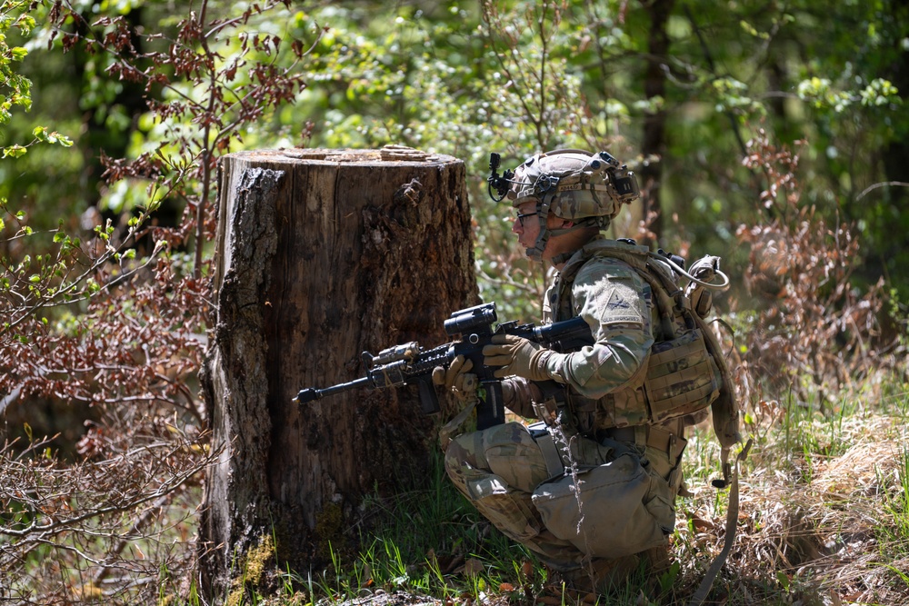 A soldier in full combat gear is kneeling in a wooded area beside a large tree stump, aiming a rifle in a tactical position. The soldier is wearing MultiCam (OCP) camouflage uniform with a tactical vest, helmet with mounted device, and backpack. The setting appears to be a training exercise or field operation in a forested environment.