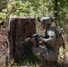 A soldier in full combat gear is kneeling in a wooded area beside a large tree stump, aiming a rifle in a tactical position. The soldier is wearing MultiCam (OCP) camouflage uniform with a tactical vest, helmet with mounted device, and backpack. The setting appears to be a training exercise or field operation in a forested environment.