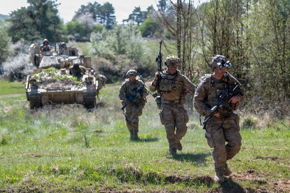 The image shows a group of military soldiers in full combat gear advancing on foot across an open grassy field. Behind them, an armored fighting vehicle (likely a Bradley IFV or similar tracked vehicle) follows in support. The scene appears to be a military training exercise or field operation, likely involving a Western military force based on equipment and uniforms.