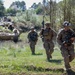 The image shows a group of military soldiers in full combat gear advancing on foot across an open grassy field. Behind them, an armored fighting vehicle (likely a Bradley IFV or similar tracked vehicle) follows in support. The scene appears to be a military training exercise or field operation, likely involving a Western military force based on equipment and uniforms.