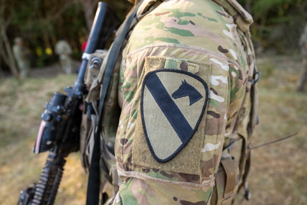 Close-up photograph of a soldier's left shoulder displaying a unit patch on a MultiCam/OCP camouflage uniform. The patch is identifiable as the 1st Cavalry Division patch, featuring a diagonal stripe and horse head silhouette on a shield-shaped background. The soldier is carrying a weapon and tactical gear in an outdoor environment.