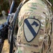 Close-up photograph of a soldier's left shoulder displaying a unit patch on a MultiCam/OCP camouflage uniform. The patch is identifiable as the 1st Cavalry Division patch, featuring a diagonal stripe and horse head silhouette on a shield-shaped background. The soldier is carrying a weapon and tactical gear in an outdoor environment.