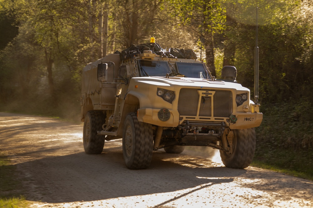 A tan/desert-colored military tactical vehicle is traveling along a narrow tree-lined gravel road, kicking up dust in warm golden sunlight. The vehicle bears the marking 'HHC7' on its front bumper and is equipped with antennas, a rooftop amber beacon, and various mounted equipment. The setting appears to be a wooded training area or rural road during late afternoon or early morning light.