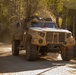 A tan/desert-colored military tactical vehicle is traveling along a narrow tree-lined gravel road, kicking up dust in warm golden sunlight. The vehicle bears the marking 'HHC7' on its front bumper and is equipped with antennas, a rooftop amber beacon, and various mounted equipment. The setting appears to be a wooded training area or rural road during late afternoon or early morning light.